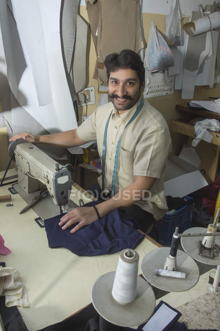 Tailleur masculin travaillant sur machine à coudre en atelier avec accessoires de couture suspendus autour — Photo de stock