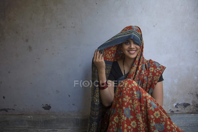 Mujer sonriente sentada en el suelo y cubriendo la cabeza con sari - foto de stock