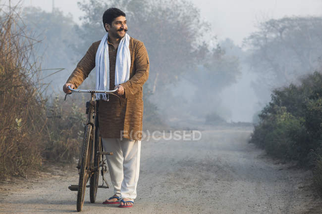 Agricoltore di sesso maschile su strada di campagna in possesso di bicicletta e guardando lontano a nebbia mattina — Foto stock
