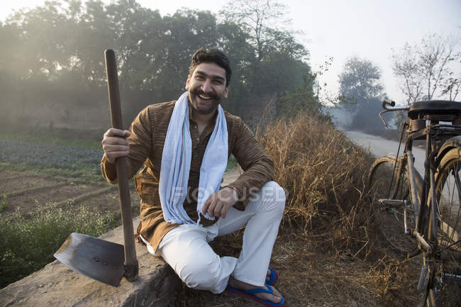 Agricultor sorridente sentado perto do campo agrícola e segurando pá — Fotografia de Stock