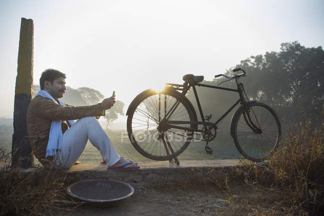 Фермер, що сидить біля сільськогосподарського поля з велосипедом і використовує мобільний телефон — стокове фото