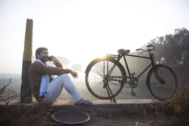 Agricultor sentado perto do campo agrícola com bicicleta e falando no celular — Fotografia de Stock