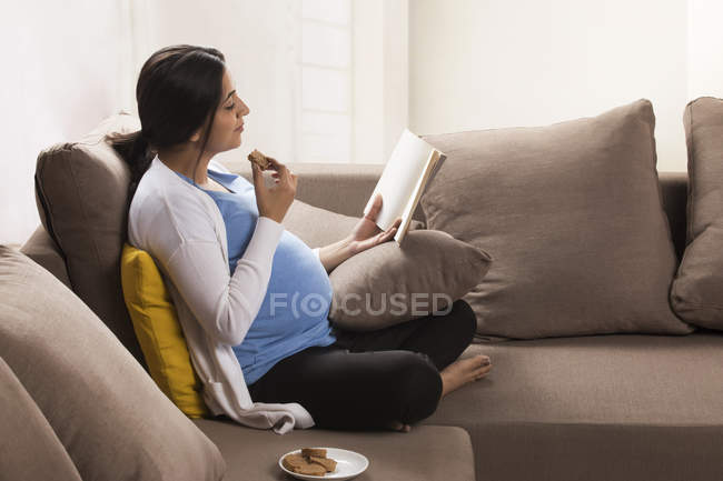 Donna incinta seduta sul divano e che mangia mentre legge un libro . — Foto stock