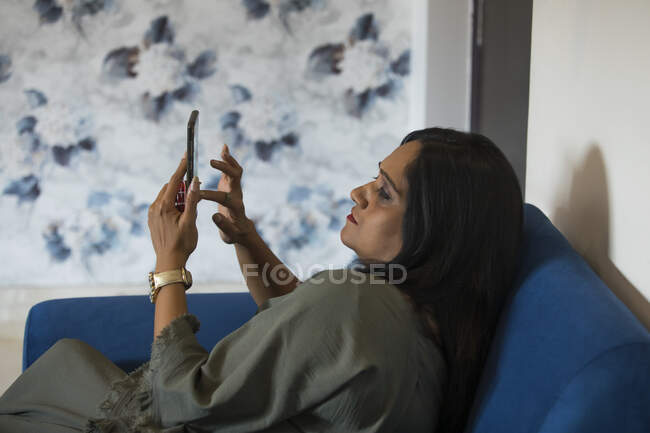 Mujer sentada en el sofá y revisando su teléfono en casa . - foto de stock