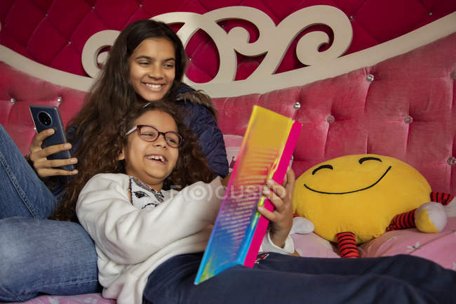Sorelle che ridono mentre leggono un libro insieme in camera da letto a casa . — Foto stock