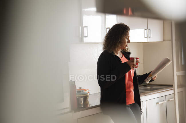 Mujer tomando café mientras lee un periódico en la cocina de su casa . - foto de stock