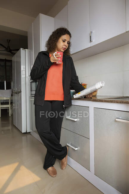 Frau trinkt Kaffee, während sie zu Hause in der Küche Zeitung liest. — Stockfoto