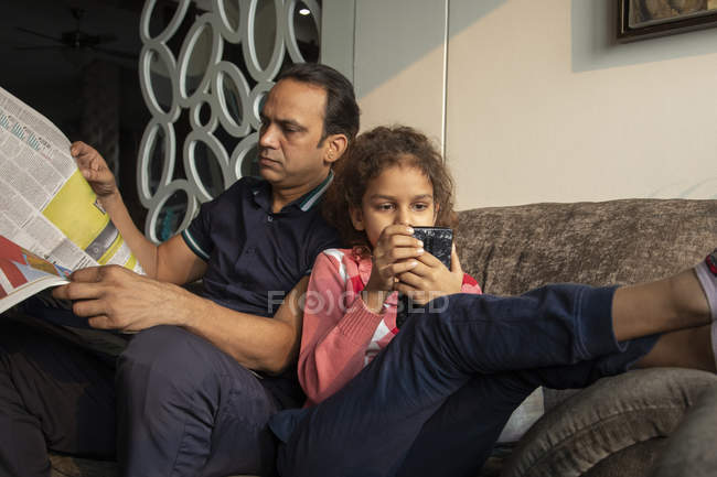 Young girl spending time on the phone while the father reads the newspaper at home. — Stock Photo
