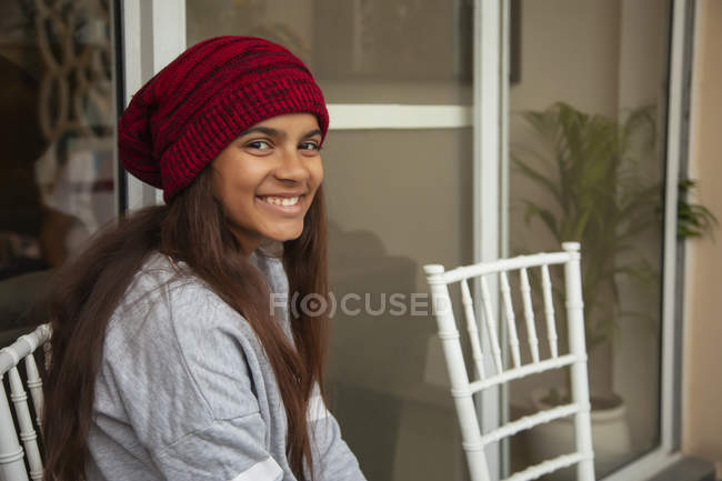 Giovane ragazza con un berretto sorridente mentre seduto sulla sedia sul balcone a casa
. — Foto stock