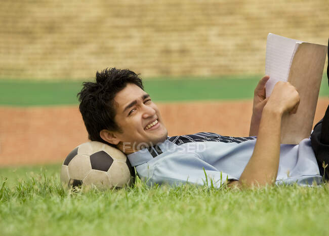 Menino deitado na bola de futebol e lendo no jardim de uma escola — Fotografia de Stock