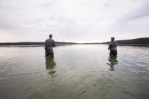 Vista trasera de dos hombres en vadeadores son la pesca con mosca en un lago al amanecer . - foto de stock