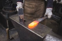 Um ferreiro martela um pedaço de metal quente em uma bigorna em uma oficina . — Fotografia de Stock