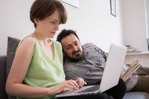 Young woman on the computer in the living room with boyfriend laying on the couch. — Stock Photo