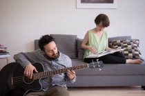 A young man is rehearsing on his bass guitar while his girlfriend is reading a book in the living room. — Stock Photo