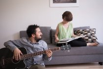 Ein junger Mann probt auf seiner Bassgitarre, während seine Freundin im Wohnzimmer ein Buch liest. — Stockfoto