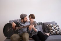 Ein junger Mann probt auf seiner Bassgitarre im Wohnzimmer, während seine Freundin ihn von der Couch aus bewundert. — Stockfoto