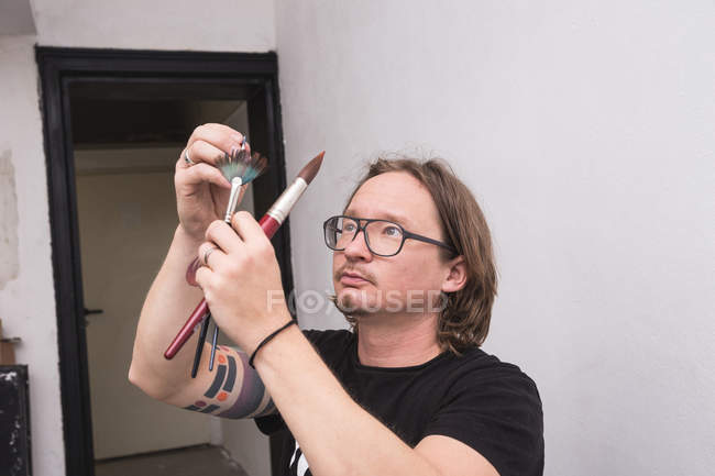 Portrait of male artist in his workshop and looking on brushes  in hands — Stock Photo