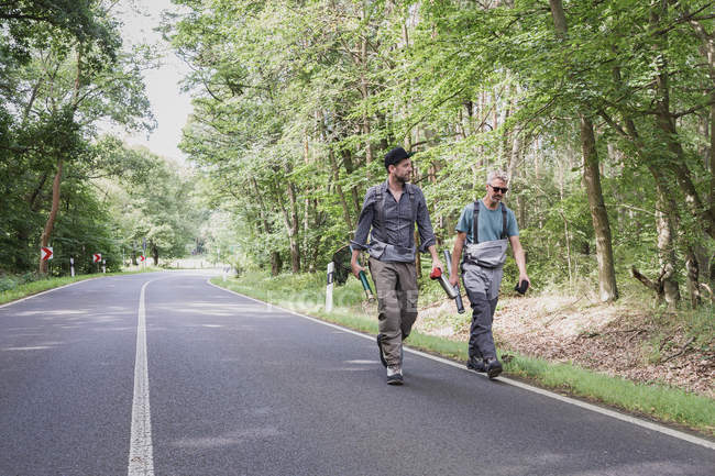 Two men with fly fishing equipment in forest area walking on road — Stock Photo