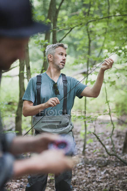 Un hombre en waders está preparando la línea para la pesca con mosca mientras que otro está preparando su caña de pescar . - foto de stock