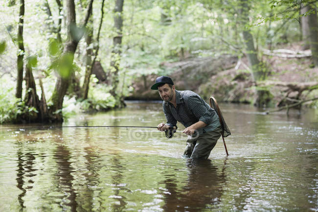 Un homme patient dans les échassiers pêche à la mouche sur la rivière dans la zone forestière . — Photo de stock