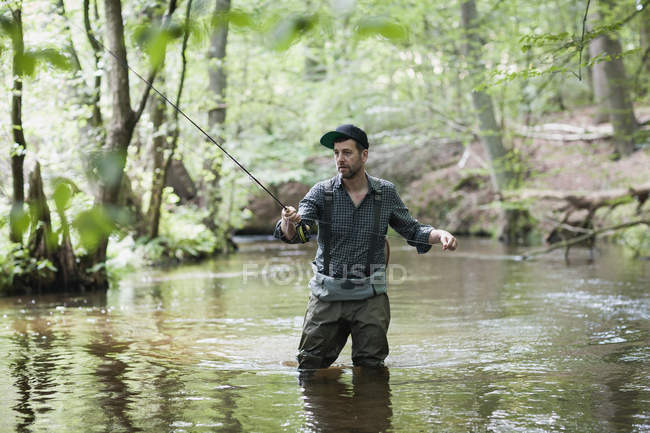 Un uomo paziente in trampolieri è pesca a mosca sul fiume nella zona della foresta . — Foto stock