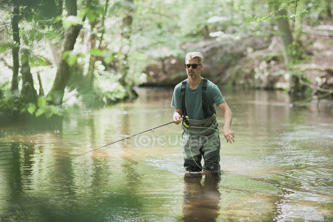 Um homem paciente em waders é pesca com mosca no rio na área florestal . — Fotografia de Stock