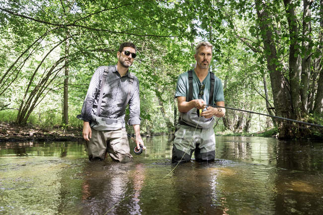 Due uomini in trampolieri sono pesca a mosca sul fiume nella zona forestale . — Foto stock