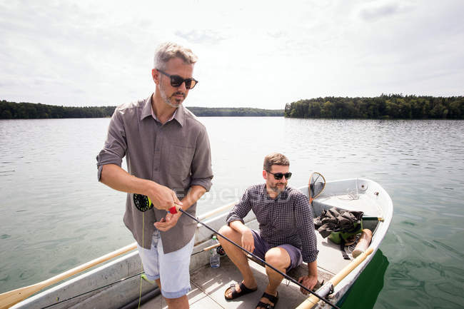 Um homem está remando um barco a remo, enquanto seu amigo é pesca com mosca de um barco no lago . — Fotografia de Stock