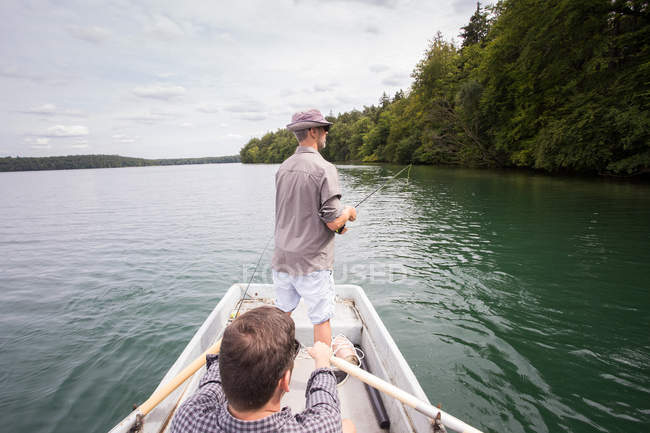 Un hombre está remando un bote de remos mientras su amigo está pescando con mosca desde un barco en el lago . - foto de stock