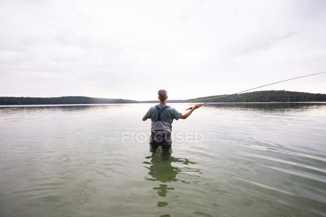 Rear view of man in waders is fly fishing in a lake. — Stock Photo