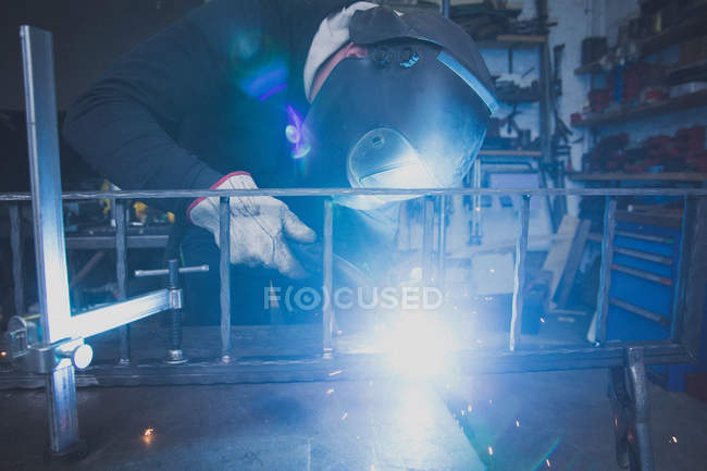 Un fabbro indossa un equipaggiamento di sicurezza e sta saldando una costruzione metallica nell'officina di un fabbro
. — Foto stock