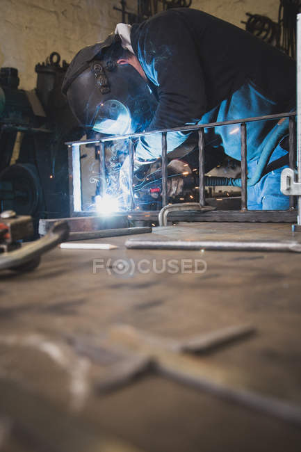 Un fabbro indossa un equipaggiamento di sicurezza e sta saldando una costruzione metallica nell'officina di un fabbro
. — Foto stock