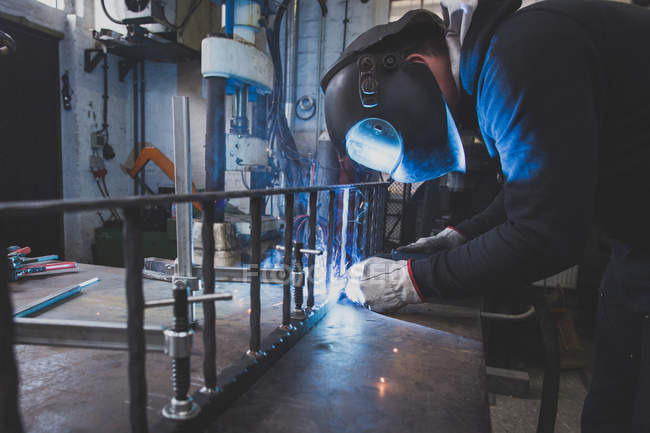 Кузнец носит защитное снаряжение и сваривает металлоконструкцию в металлообрабатывающей мастерской
. — стоковое фото