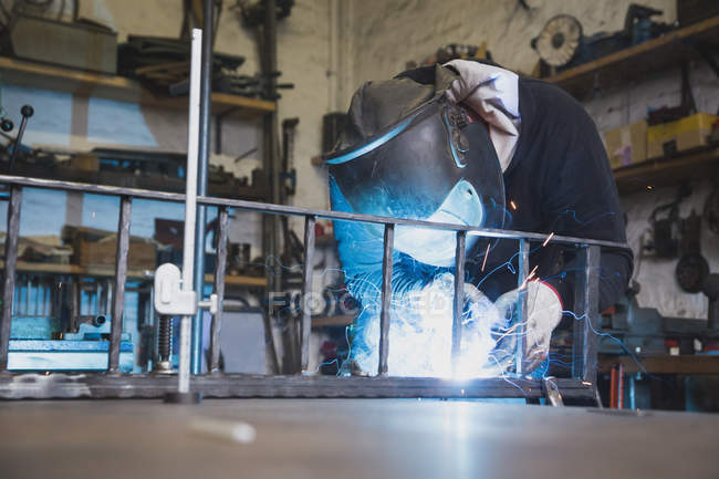 Кузнец носит защитное снаряжение и сваривает металлоконструкцию в металлообрабатывающей мастерской . — стоковое фото