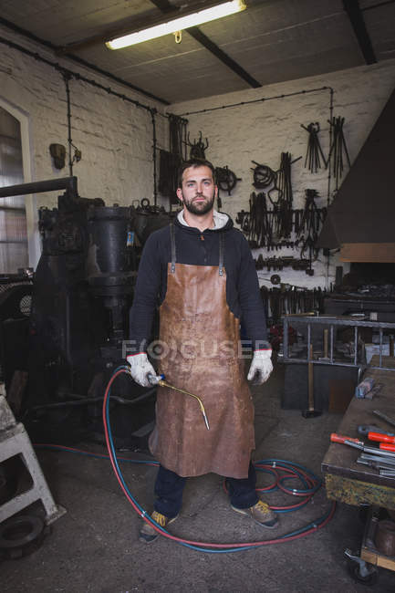 Un herrero en un delantal de cuero y con una antorcha de corte es retratado en su taller
. - foto de stock