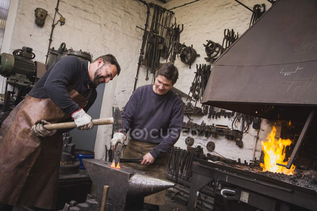 Dos herreros martillan un trozo de metal en un yunque en un taller . - foto de stock