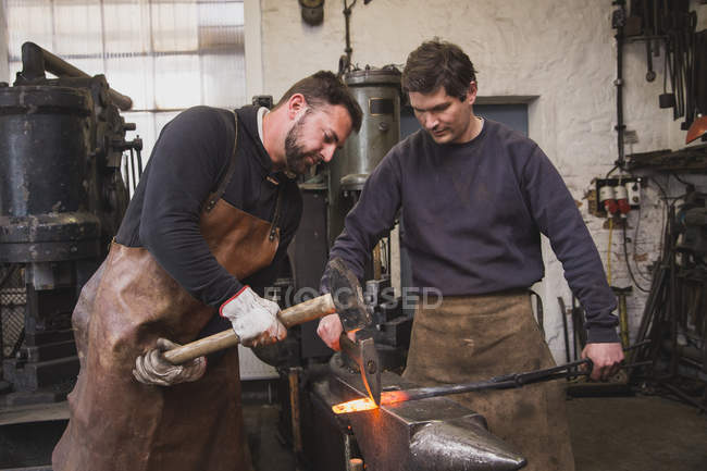 Two blacksmiths hammer a piece of metal on an anvil in a workshop. — Stock Photo
