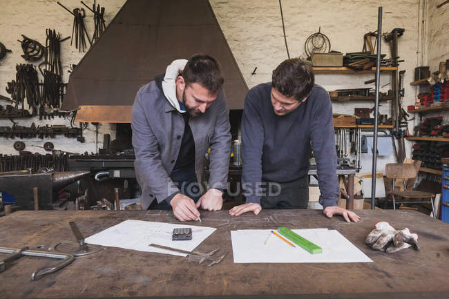 Two blacksmiths are taking measures, do calculations and schedule a days work in a blacksmith's workshop. — Stock Photo