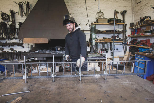 Un forgeron souriant porte un équipement de sécurité et est sur le point de souder une construction métallique dans un atelier de forgeron . — Photo de stock