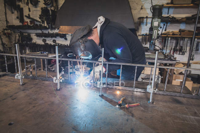 Un fabbro indossa un equipaggiamento di sicurezza e sta saldando una costruzione metallica nell'officina di un fabbro . — Foto stock