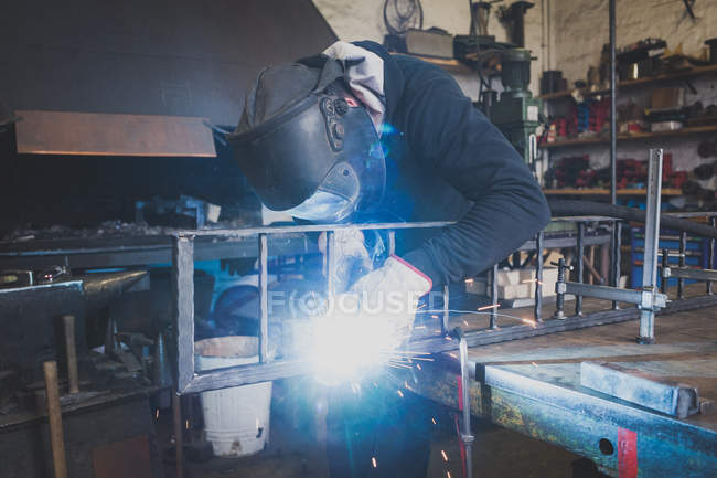 Um ferreiro usa engrenagem de segurança e está soldando uma construção de metal na oficina de um metaleiro . — Fotografia de Stock