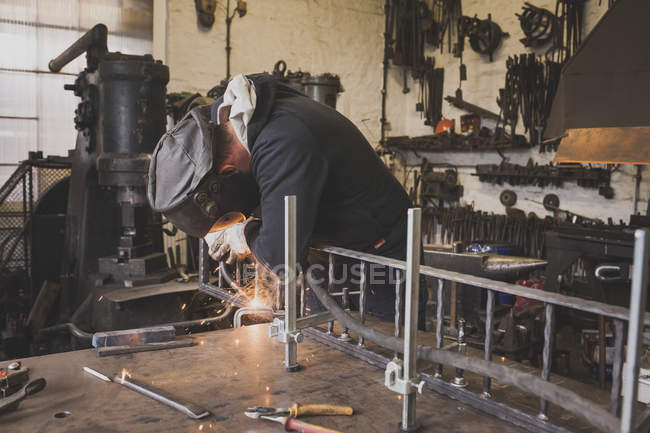 Ein Schmied trägt Schutzausrüstung und schweißt eine Metallkonstruktion in einer Schlosserei. — Stockfoto