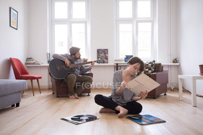 Un giovane uomo sta provando il suo basso mentre la ragazza sta controllando i dischi in vinile in salotto . — Foto stock