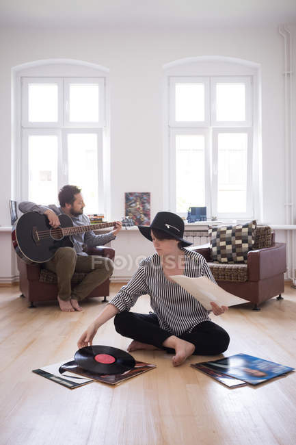 A young man is rehearsing on his bass guitar while the girlfiend is checking out vinyl records in the living room. — Stock Photo