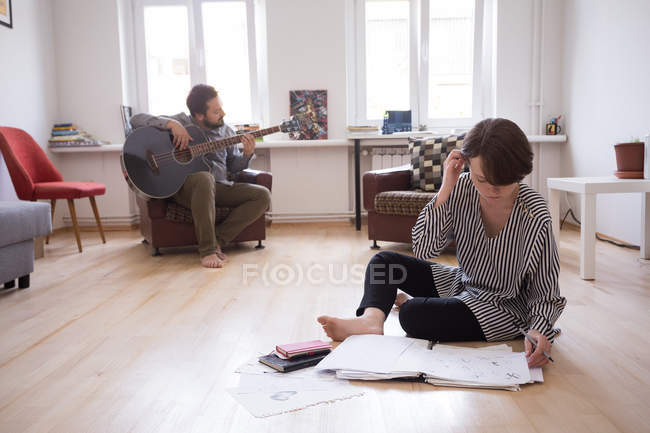 Ein junger Mann probt auf seiner Bassgitarre, während die Freundin im Wohnzimmer Zeichnungen anfertigt. — Stockfoto