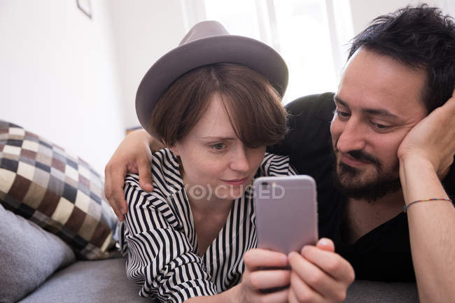 Una joven está revisando su teléfono inteligente mientras su novio se relaja con ella en el sofá . - foto de stock
