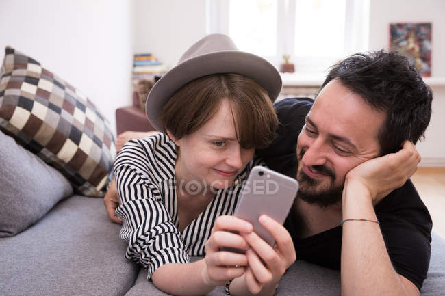 Um jovem casal está fazendo selfies com um telefone inteligente enquanto relaxa no sofá durante o fim de semana . — Fotografia de Stock