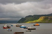 Reino Unido, Escocia, Highland, Isla de Skye, Portree Harbor con barcos y acantilados verdes en el clima malhumorado - foto de stock