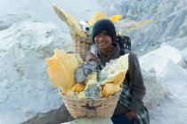 JAVA, INDONÉSIA - 18 DE JUNHO DE 2018: Mineração de enxofre no vulcão Ijen, homem carregando enxofre em cestas por cratera, olhando para a câmera, sorrindo — Fotografia de Stock