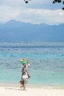 Indonesia, Nusa Tenggara Barat, Lombok Utara, mujer llevando cesta en la cabeza en la playa - foto de stock